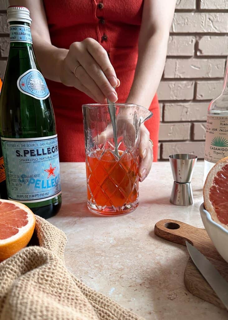 stiring an aperol paloma cocktail in a glass pitcher