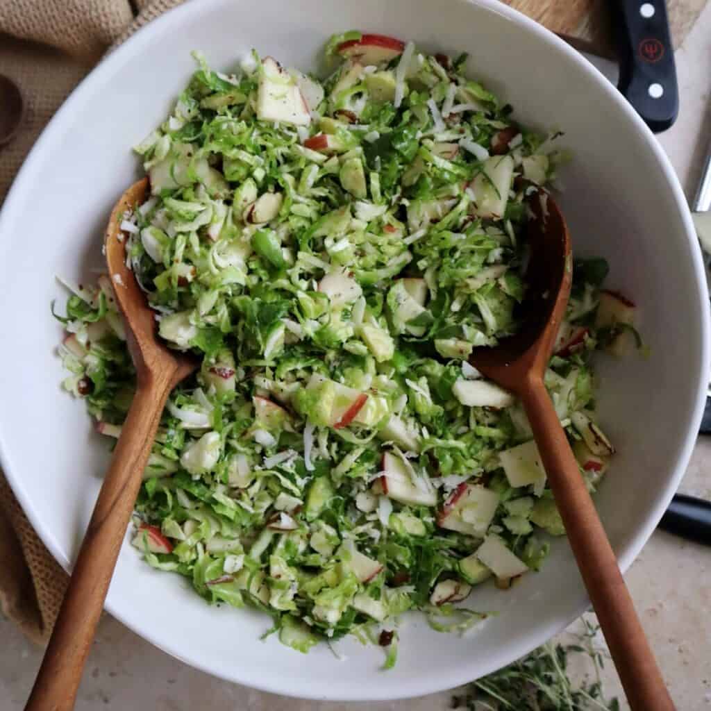 shaved brussels sprouts salad with apple manchego and almond in white bowl with wooden salad spoons