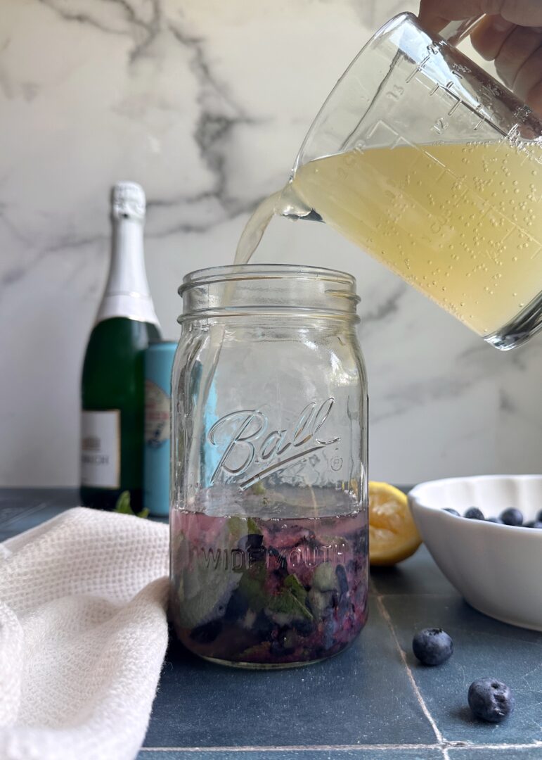 adding ginger beer to smashed blueberries in a glass jar