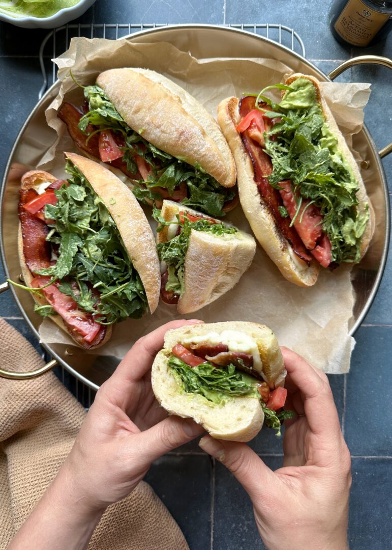 half of a green goddess caprese melt held over a tray of other sandwiches