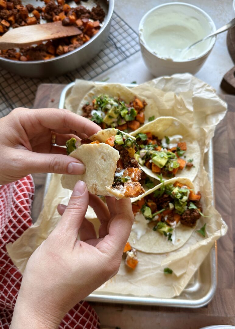 holding a chorizo sweet potato taco with a tray of tacos underneath