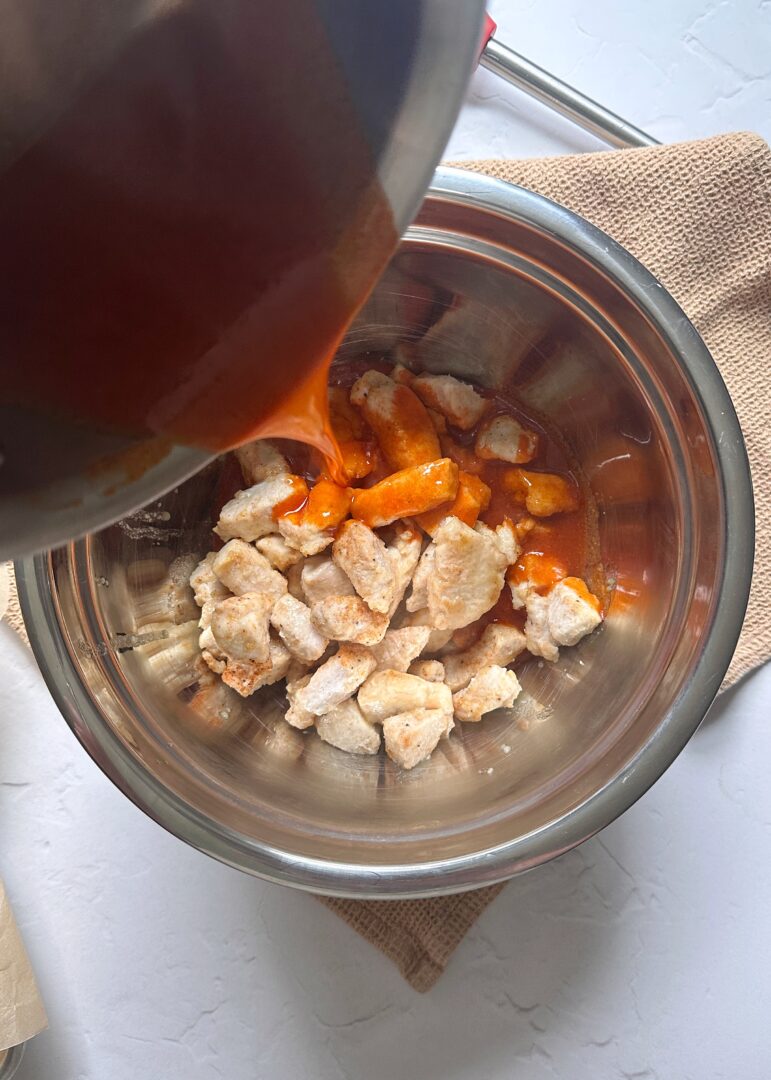 pouring buffalo sauce over baked chicken wings in a mixing bowl