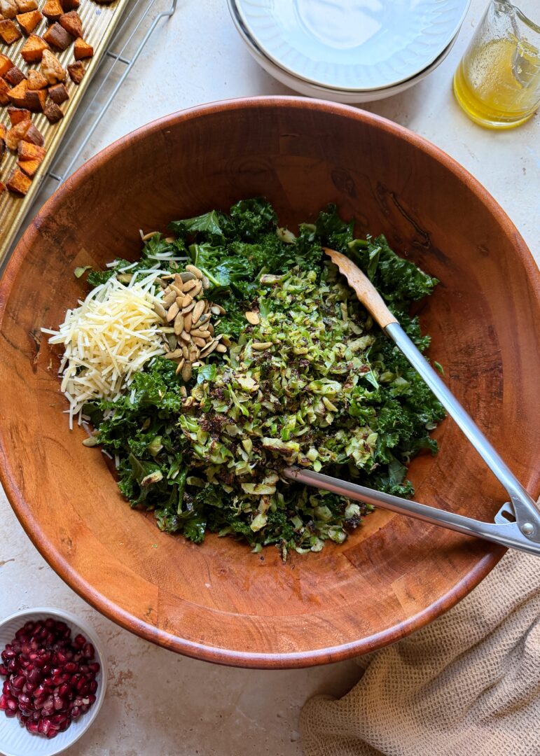 mixing roasted sprouts parmesan and pumpkin seeds in a kale salad with tongs